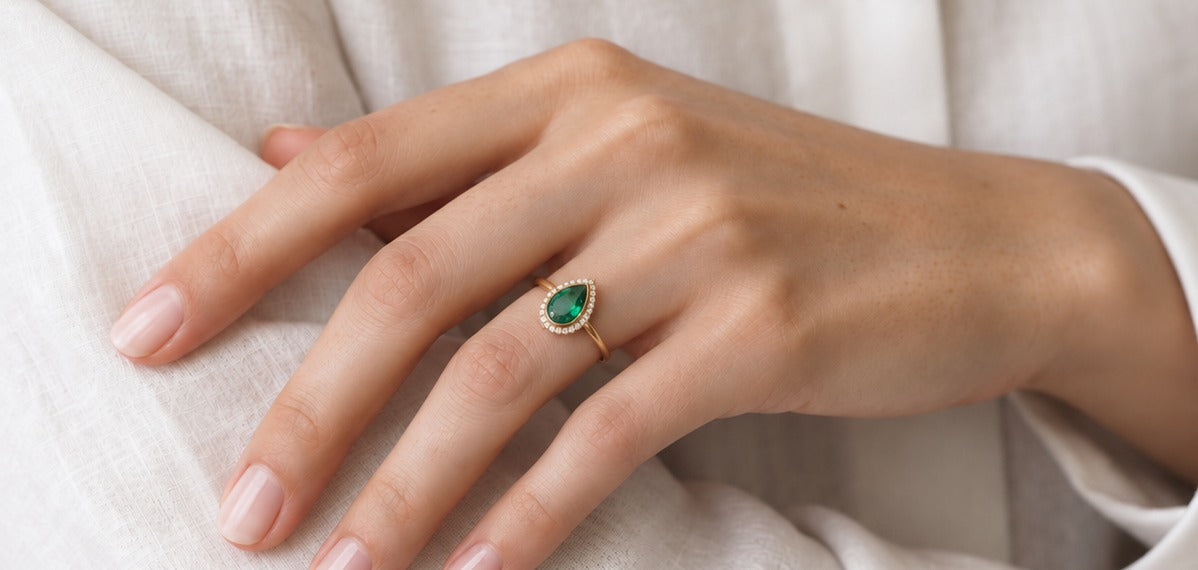Bague Ocelle portée à la main sur une chemise en lin, émeraude poire et halo de diamants, or jaune